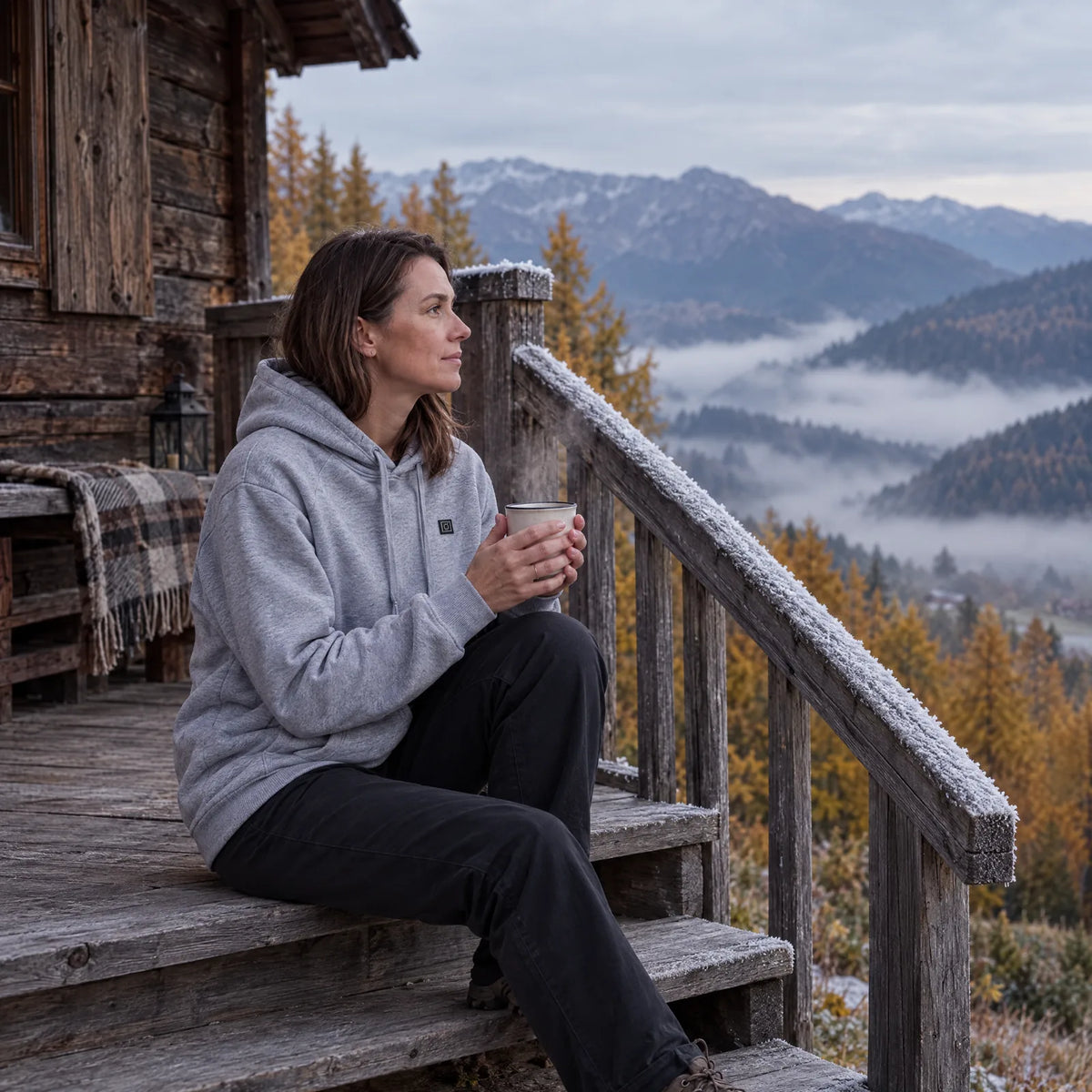 Woman in the gray heated hoodie with tea on a mountain hut porch on an autumn morning